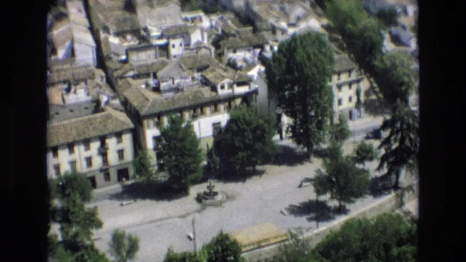 1972 Spain 8mm frame with a high-angle view across tiled rooftops, pale town buildings, trees, and a small fountain in an open square.
