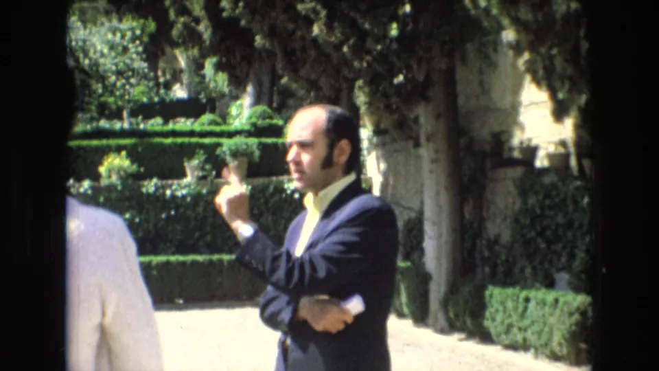 1972 Spain home-movie frame showing an adult man in a dark suit and light shirt gesturing during conversation beside manicured hedges and trees in a garden.