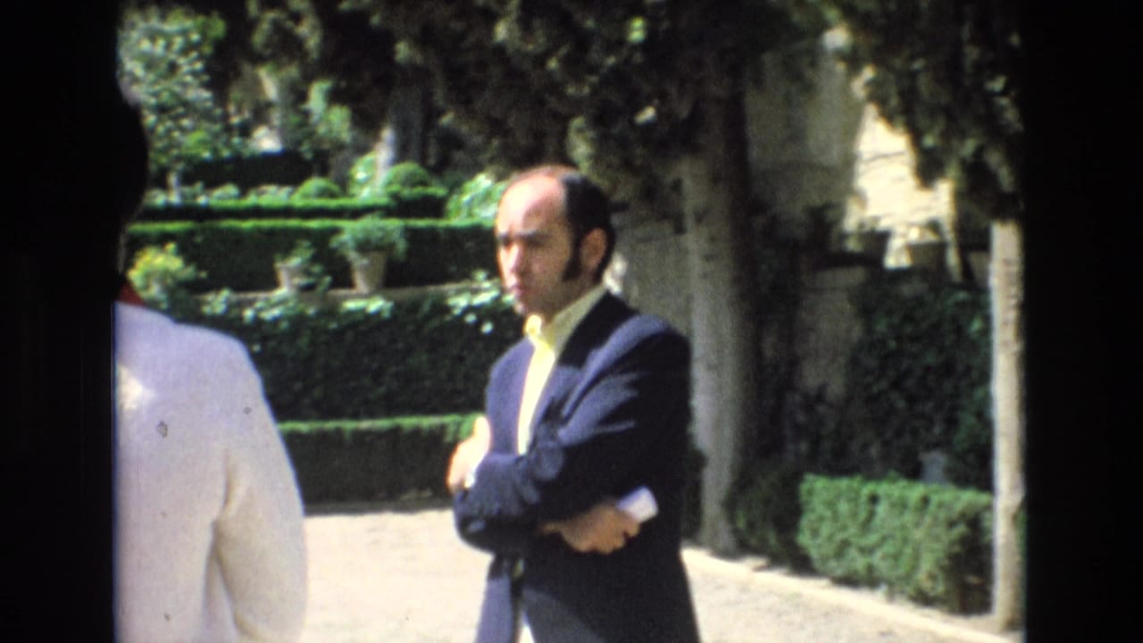 In 1972 Spain home-movie footage, a man in a dark suit and pale yellow shirt stands in a garden with trimmed hedges and trees as a companion is partly visible at left.
