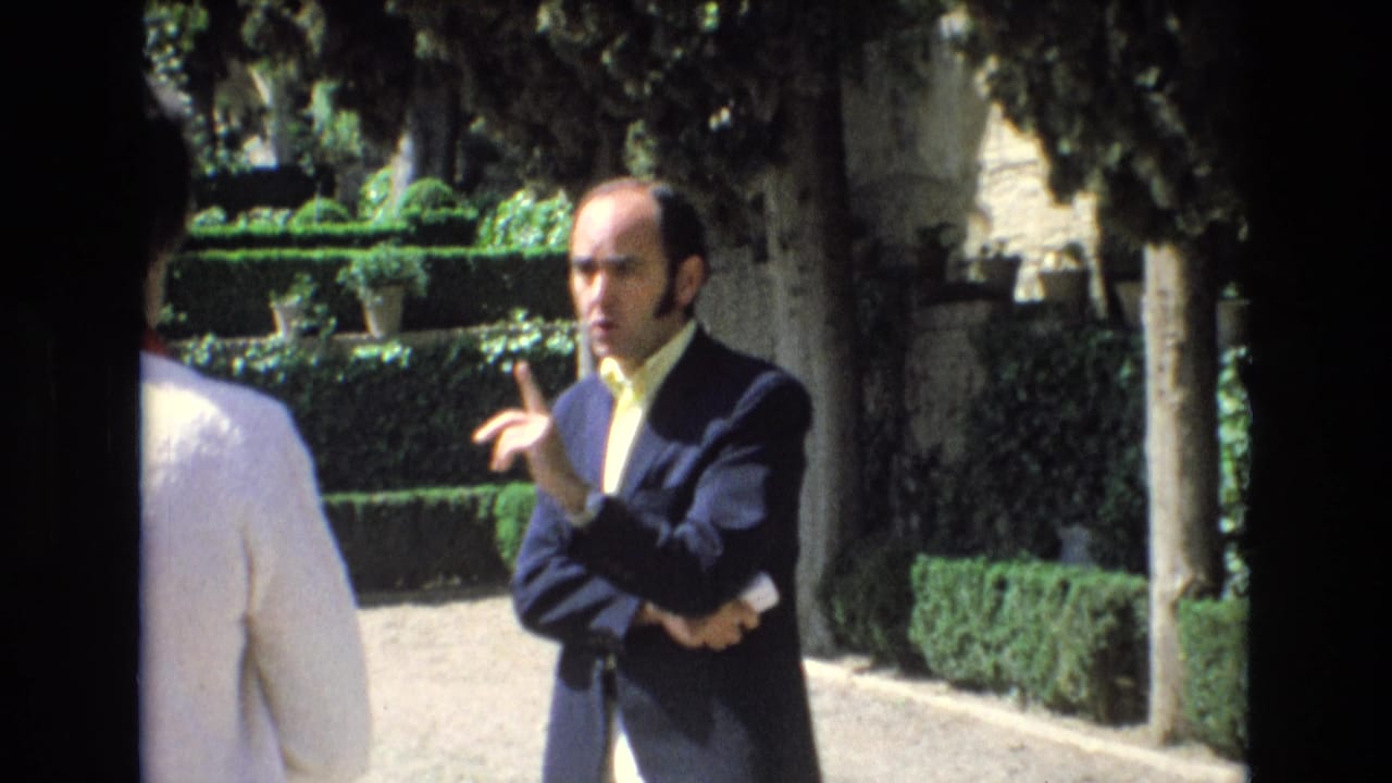 Man in a dark suit gestures while standing in a garden beside a partially visible person.