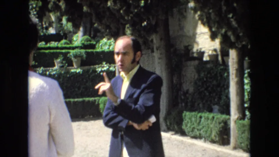 In the garden sequence, the suited man gestures with one hand while facing someone nearby, with clipped hedges, trees, and a sunlit path behind him.