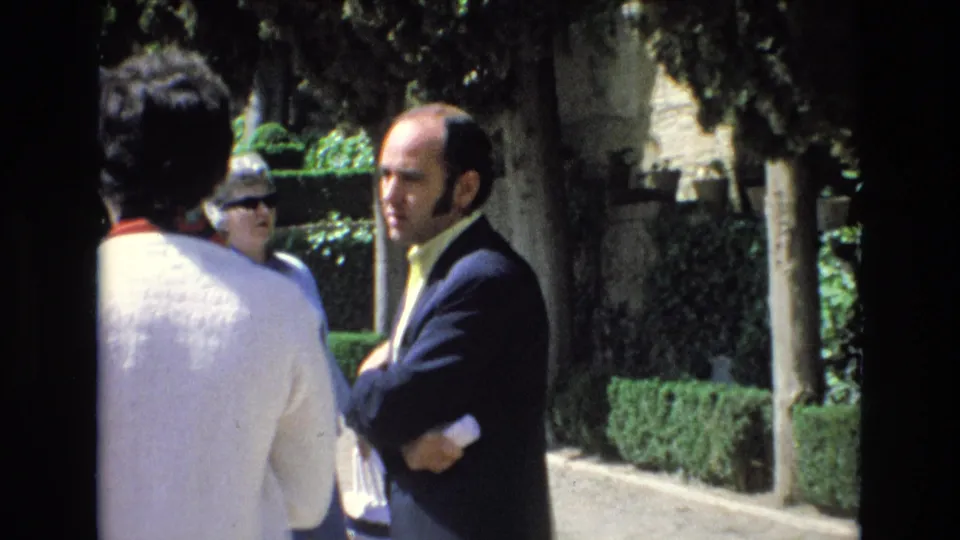 Adults appear gathered in a Spanish garden scene, with the suited man turned partly toward the camera and other people visible from behind or behind sunglasses.