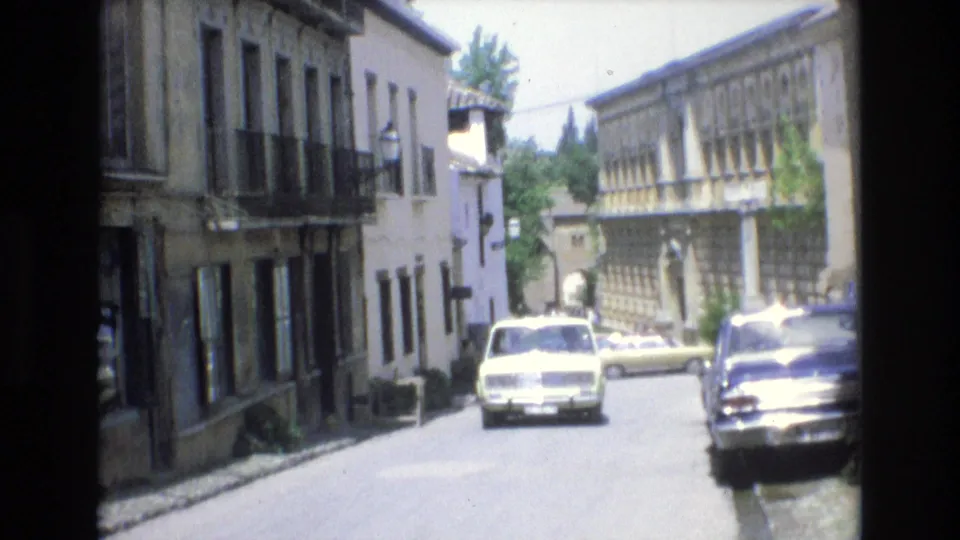 A narrow street in Spain is lined with traditional buildings and classic cars, with one light-colored car facing the camera and other vehicles parked along the curb.