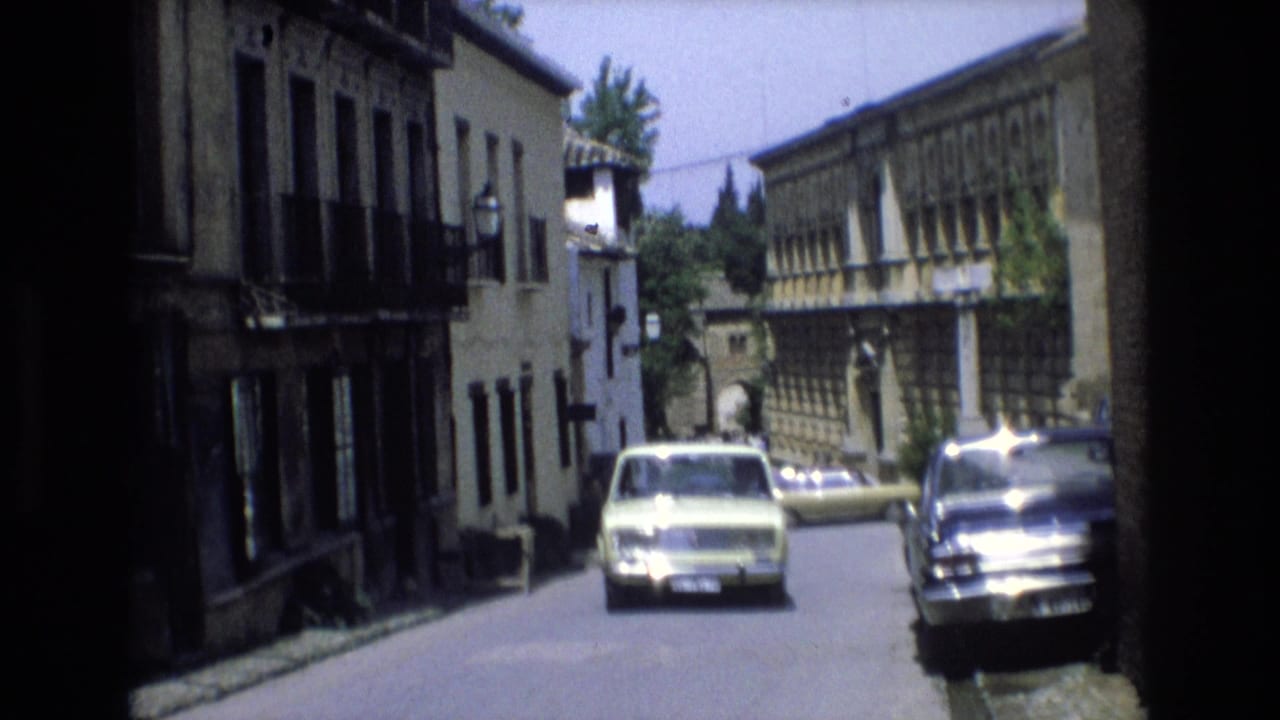 A 1972 Spain 8mm frame shows a pale classic car approaching along a narrow street lined with weathered buildings, balconies, and shuttered windows.