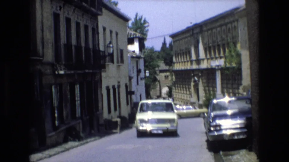 A 1972 Spain 8mm frame shows a pale classic car approaching along a narrow street lined with weathered buildings, balconies, and shuttered windows.
