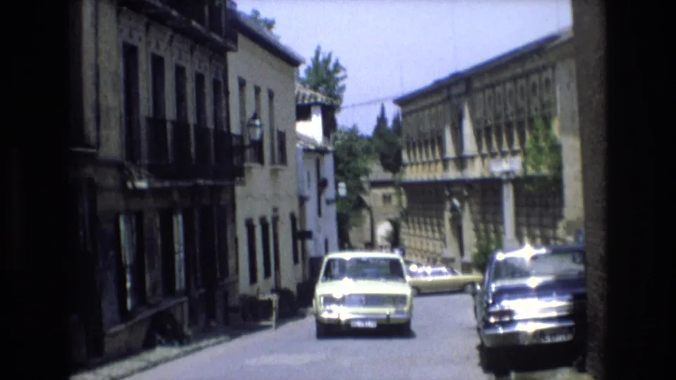 The 8mm frame holds a shaded foreground view onto a bright Spanish street where a pale car approaches between stone-fronted buildings and shuttered windows.