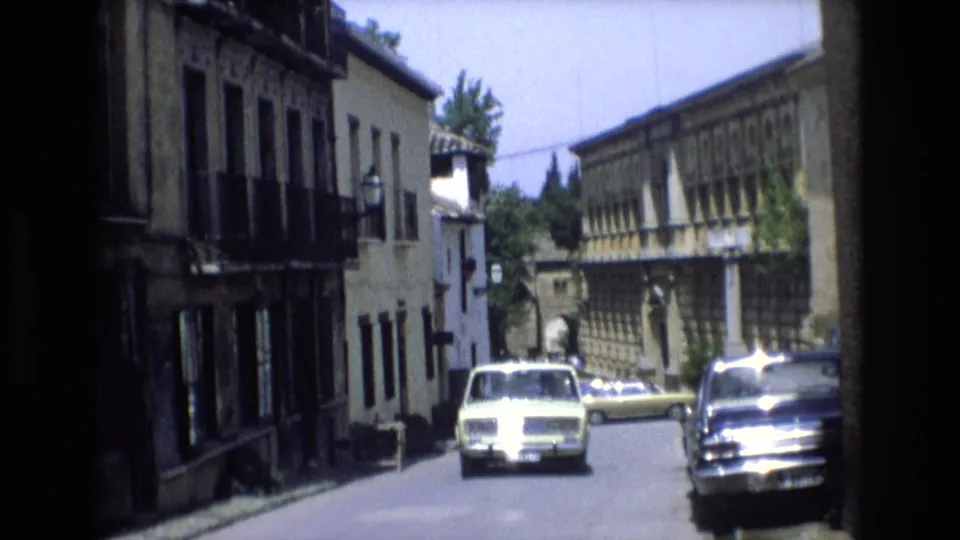 A final 1972 Spain street frame shows the pale car centered near the camera, a dark parked sedan at right, and sunlit historic facades receding down the road.