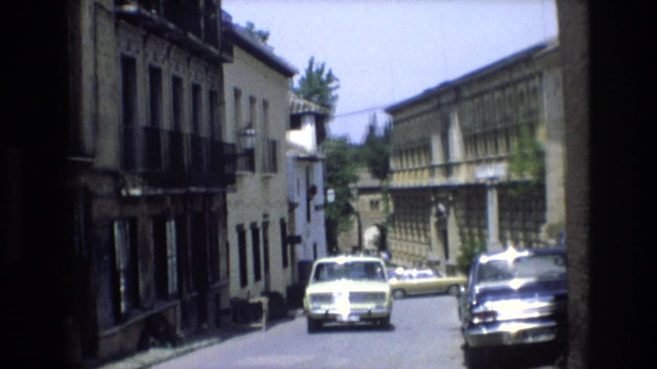 1972 Spain travel footage frame showing a narrow paved street lined with older buildings, balconies, and parked compact cars in bright daylight.