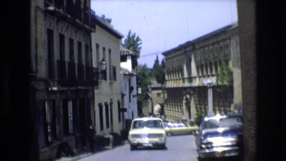 A Spain 1972 street scene shows compact cars parked along a narrow roadway between buildings with balconies and shuttered upper windows.