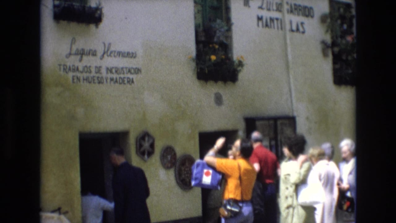 1972 Spain home-movie frame showing tourists outside a stucco craft-shop facade with Spanish signage and a woman in an orange shirt taking a photograph.