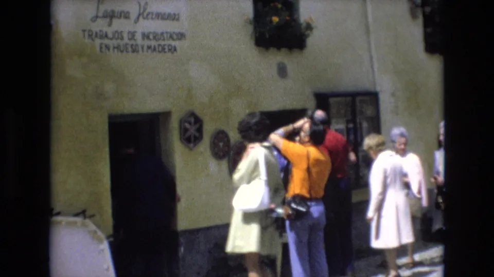 1972 Spain travel frame with visitors in period clothing near a rustic shop entrance beneath Spanish signage, including a person photographing the facade.