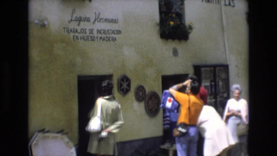 1972 Spain 8mm frame showing a tourist with a camera facing a craft-shop wall as other visitors move around the entrance.