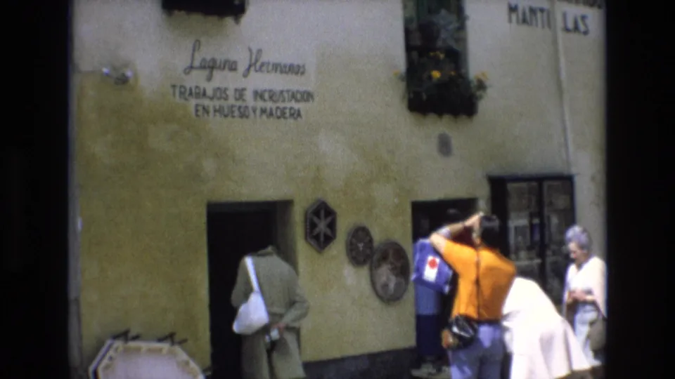 1972 Spain travel frame with tourists outside Laguna Hermanos signage, a dark doorway, wall decorations, and a handheld camera raised toward the building.
