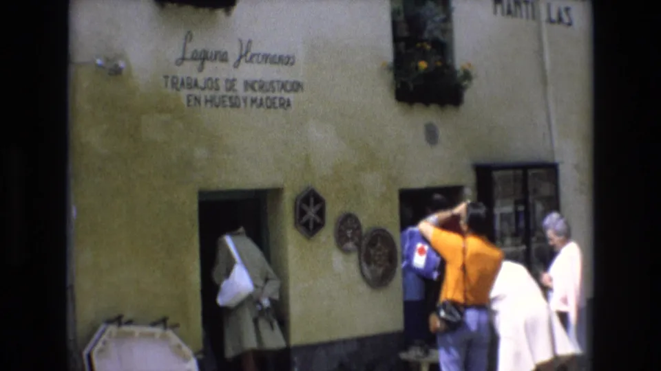 In Spain in 1972, adult tourists gather at the entrance of a stucco-fronted Laguna Hermanos workshop with a painted sign and decorative wall pieces visible.