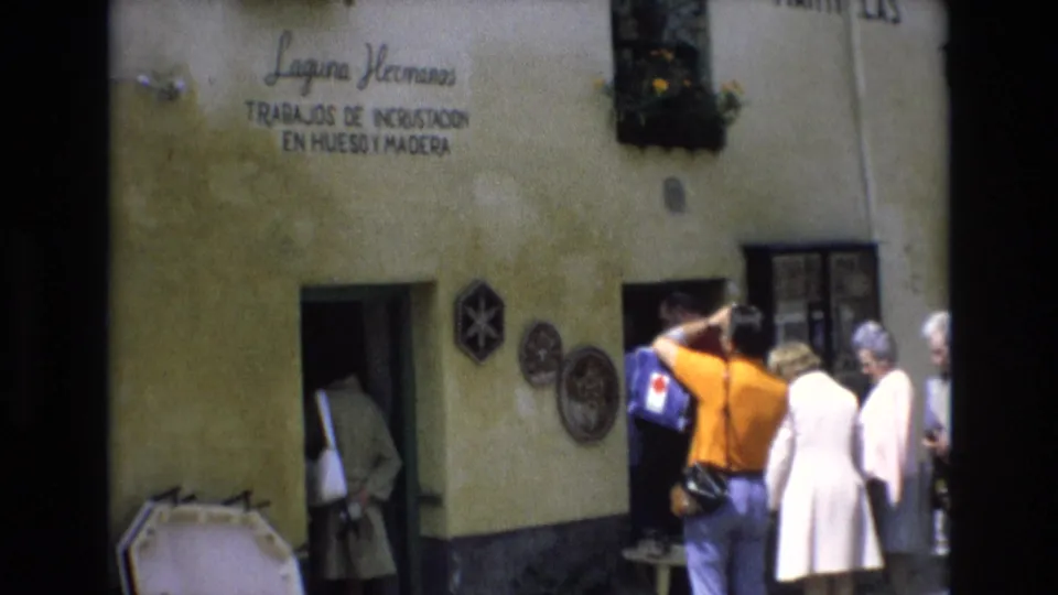 Adult visitors in 1970s clothing stand near the Laguna Hermanos doorway in Spain, with a person in an orange shirt and others in light coats facing the entrance.