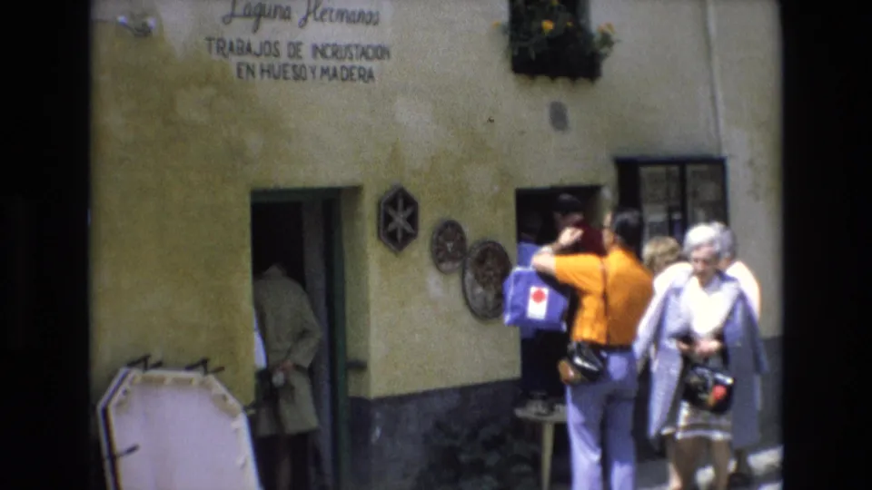 The 1972 Spain home-movie frame shows tourists with cameras and shoulder bags outside the Laguna Hermanos workshop, with the doorway and painted facade partly cropped.