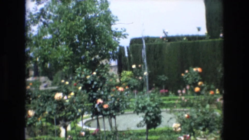 1972 Spain home-movie frame showing a circular garden fountain pool with rose bushes, clipped hedges, and a leafy tree in bright daylight.