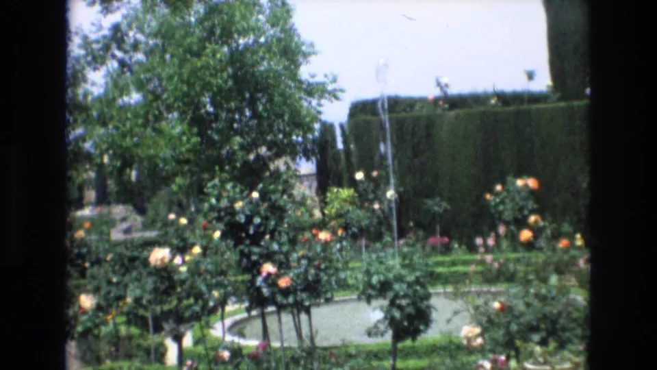 1972 Spain home-movie frame with a soft-focus view of a fountain pool, flowering rose bushes, clipped hedges, and a tree at left.