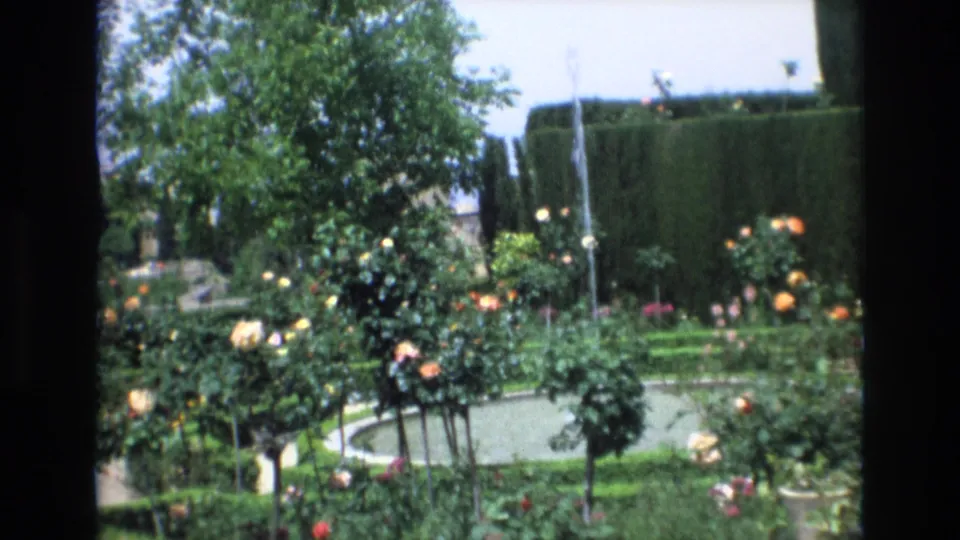 1972 Spain home-movie frame showing the circular fountain pool and surrounding rose beds, with clipped hedges and a leafy tree behind.