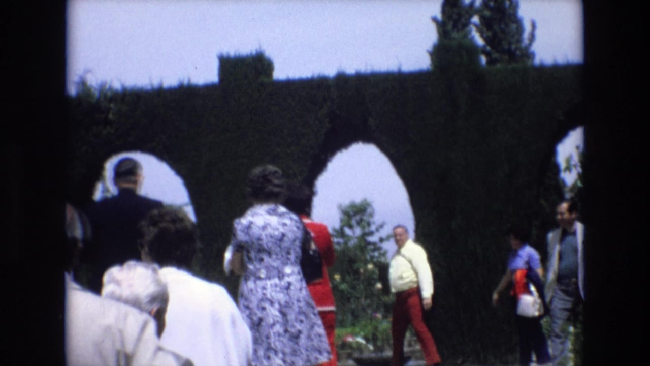 1972 Spain 8mm frame showing adult visitors moving through a formal garden with clipped hedge archways and flower beds.