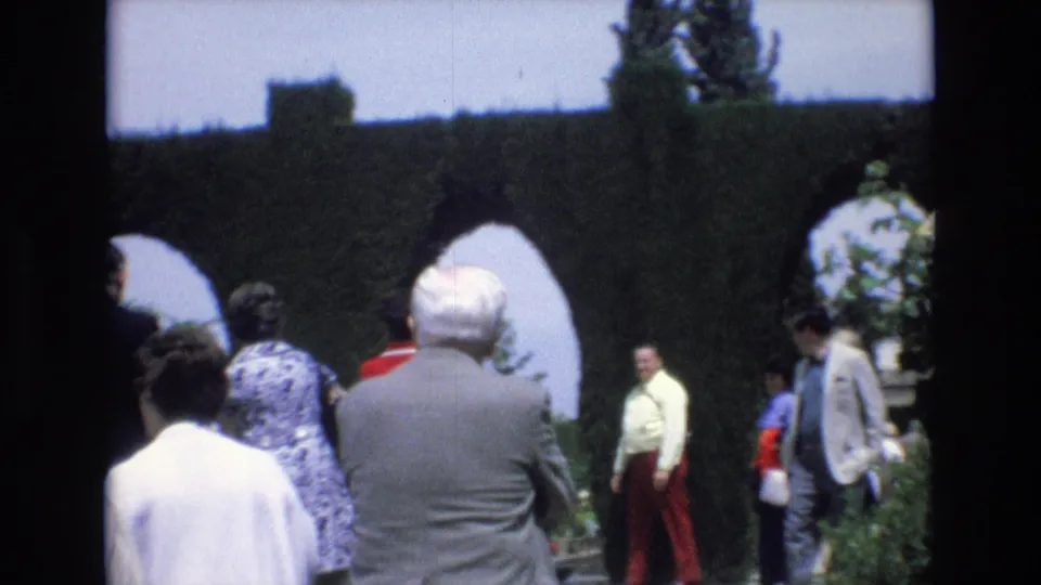1972 Spain 8mm frame with several adult visitors near arched topiary, including a man in a grey suit seen from behind.