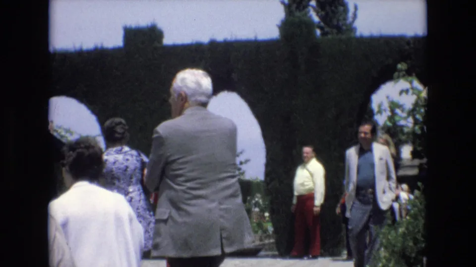 1972 Spain 8mm frame showing a grey-suited man in the foreground with other adult visitors moving through arched topiary and flower beds.