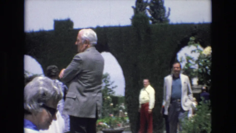 1972 Spain 8mm frame with adult visitors gathered beside clipped garden archways, including a grey-suited man and a man in a light blazer.