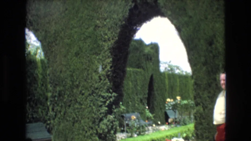 Spain 1972 garden-tour frame with a dense green hedge arch, bright opening beyond it, flower beds below, and a visitor partly visible at the edge of frame.