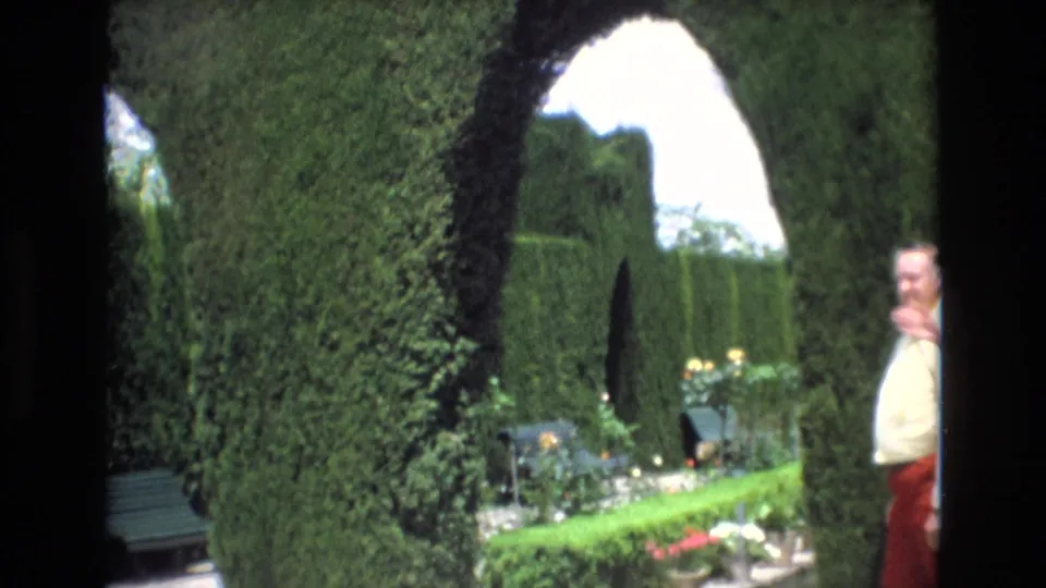 Spain 1972 home-movie frame of a formal garden with an arched hedge, low flowers, a bench, and a man in a light shirt and red trousers looking toward the camera.