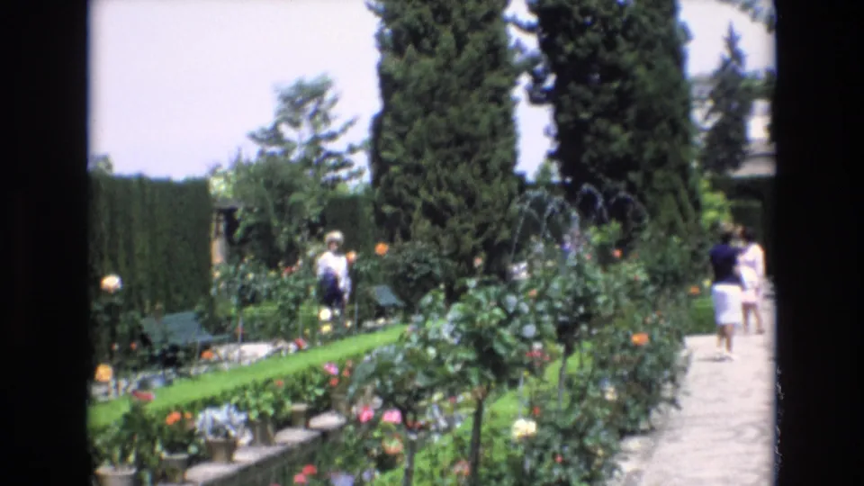 Spain 1972 garden-tour frame showing visitors walking along a paved path beside formal planting beds, tall hedges, mature trees, and a fountain area in the distance.
