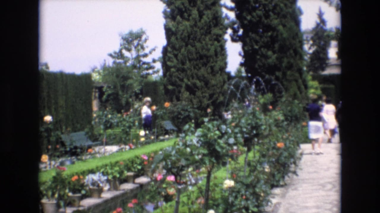 1972 Spain home-movie frame showing visitors walking through a formal garden with manicured hedges, flowering rose bushes, tall cypress trees, and a stone pathway.