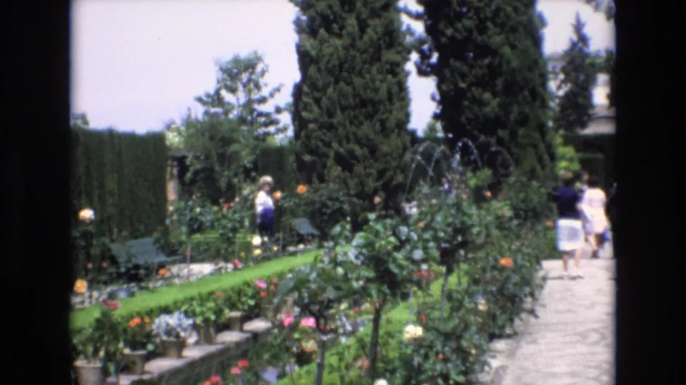 1972 Spain home-movie frame showing visitors walking through a formal garden with manicured hedges, flowering rose bushes, tall cypress trees, and a stone pathway.