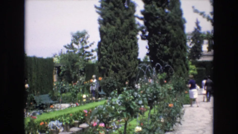 1972 Spain 8mm frame showing a visitor-shot garden scene with a stone walkway, trimmed hedges, flowering rose bushes, benches, tall cypress trees, and people in summer clothing.