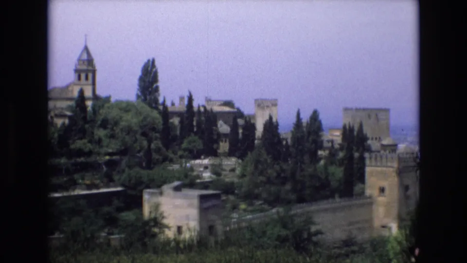 1972 Spain home-movie frame showing a distant historic stone cityscape with crenellated walls, towers, dense trees, and a church-like spire rising above the greenery.