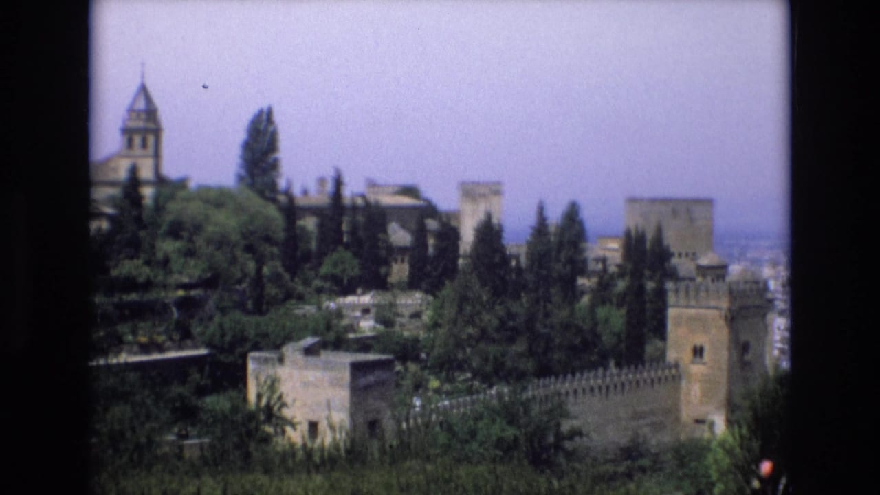 1972 Spain 8mm travel frame showing a distant historic castle complex with stone towers, crenellated walls, tall trees, and layered garden greenery.