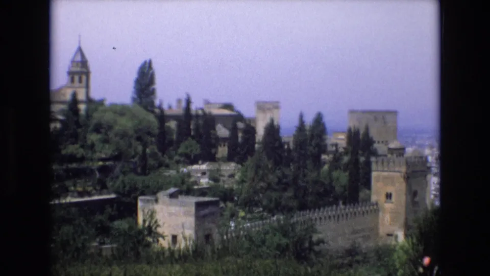 1972 Spain 8mm travel frame showing a distant historic castle complex with stone towers, crenellated walls, tall trees, and layered garden greenery.