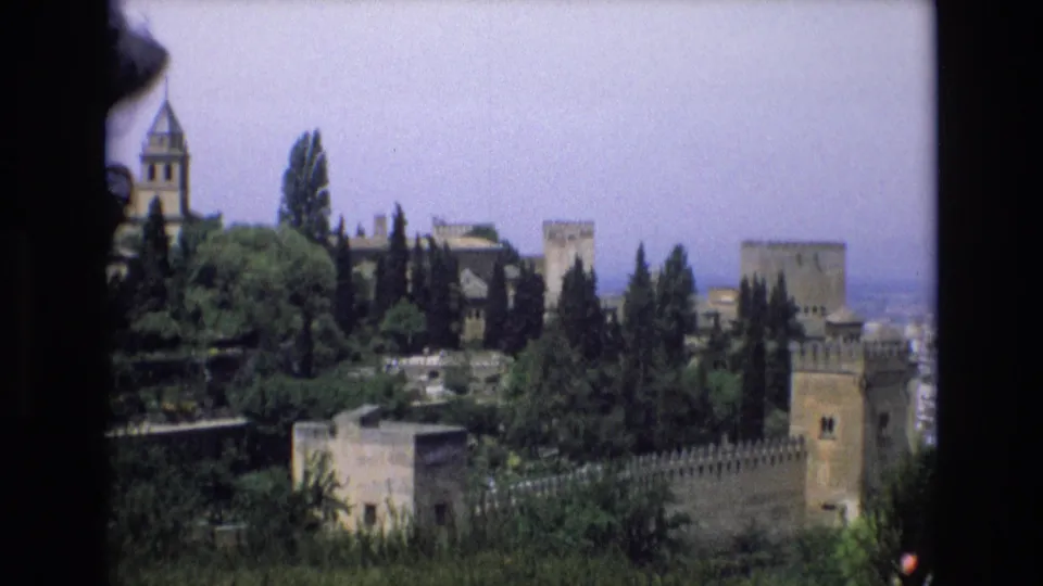1972 Spain 8mm frame with a visitor-shot view across greenery toward a castle complex, including towers, crenellated walls, and a prominent vertical tower at left.