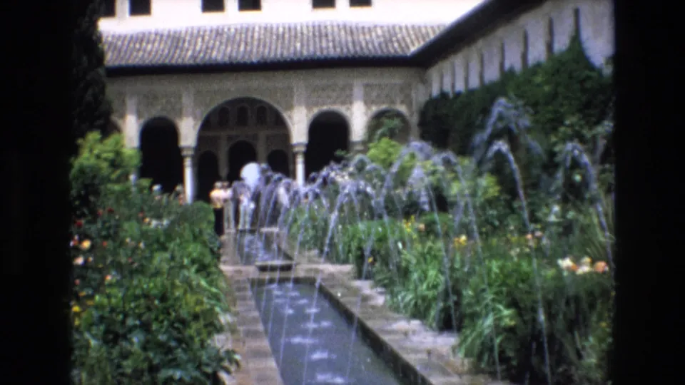 1972 Spain 8mm frame showing a long courtyard fountain, arcing water jets, flowerbeds, visitors, tiled roofing, and arched colonnades.