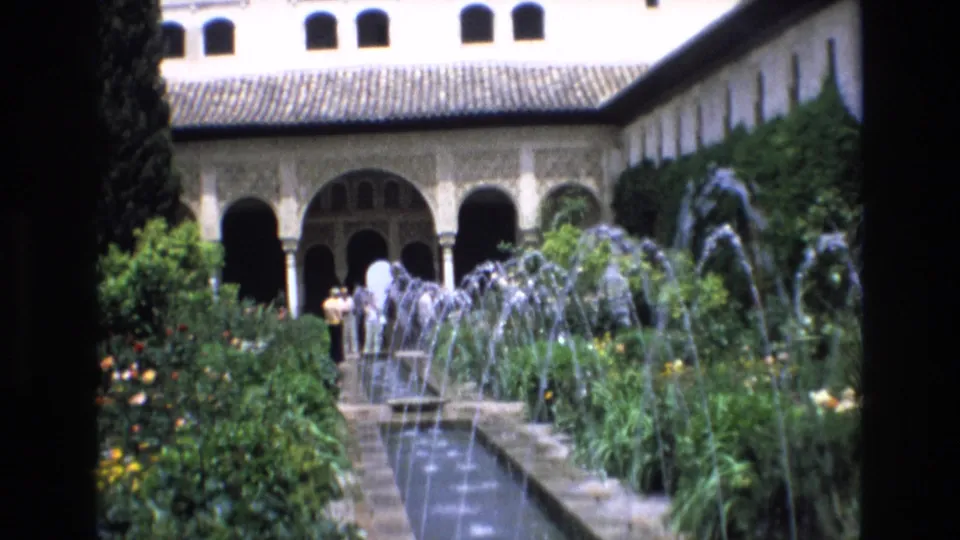 1972 Spain 8mm frame showing fountain jets, dense planting, a tiled-roof courtyard building, arched openings, and distant visitors.