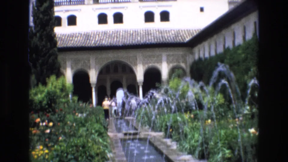 1972 Spain vacation footage showing a formal courtyard garden with water jets, greenery, arched architecture, tiled roofs, and distant visitors.