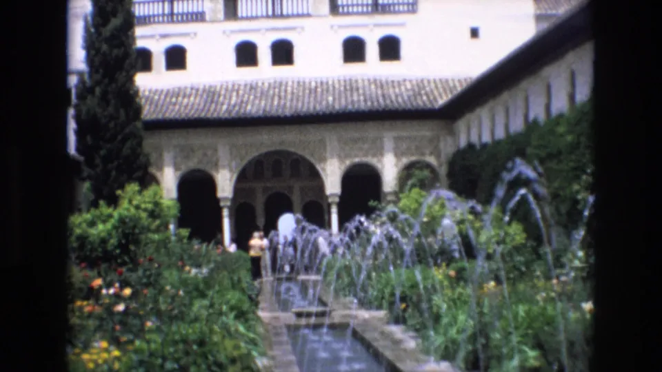 A handheld 8mm view in Spain shows a courtyard fountain garden, arched walkway, tiled roof, dense greenery, and distant tourists.
