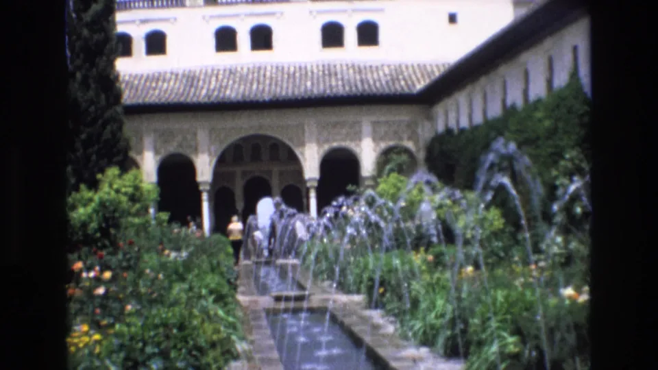 1972 Spanish travel footage captures fountains, flowering shrubs, a cypress-like tree, arched colonnades, and distant visitors in a historic garden courtyard.