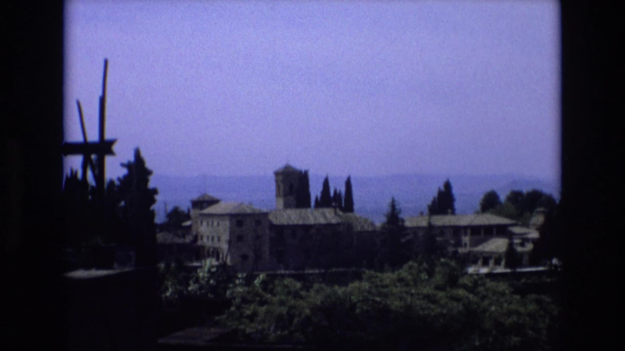 1972 Spain home-movie frame showing a distant stone building complex with a square tower, tiled roofs, cypress trees, hazy hills, and dark framing at the left edge.