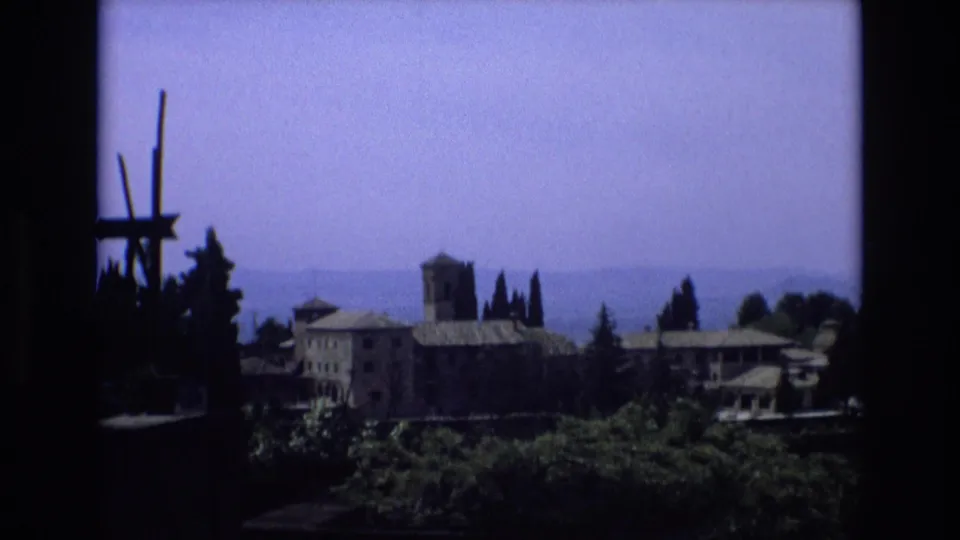 1972 Spain home-movie frame showing a distant stone building complex with a square tower, tiled roofs, cypress trees, hazy hills, and dark framing at the left edge.