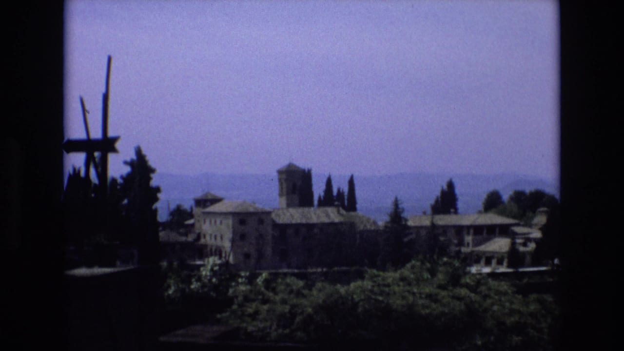 1972 Spain travel-style 8mm frame with a stone complex, prominent tower, cypress trees, blue haze over distant hills, and dark window-like borders.