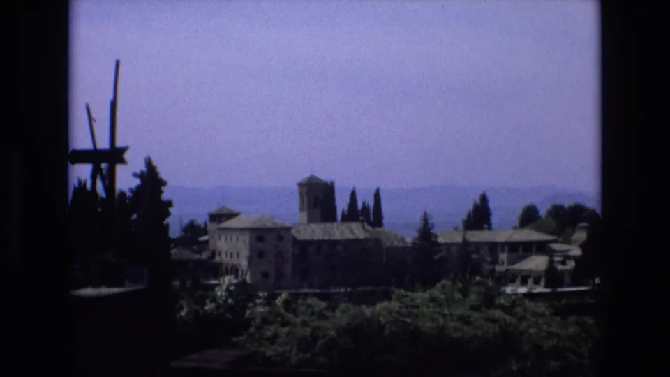 1972 Spain travel-style 8mm frame with a stone complex, prominent tower, cypress trees, blue haze over distant hills, and dark window-like borders.