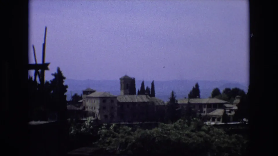 1972 Spain 8mm frame showing a stone building group with a tower and tiled roofs, partly obscured by trees and framed by a dark silhouette on the left.