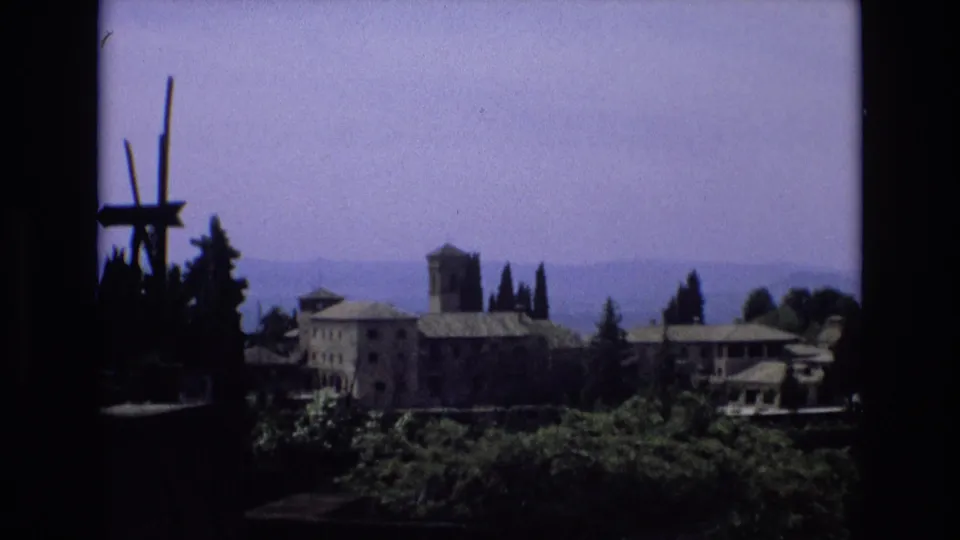 1972 Spain home-movie frame of a distant stone building complex with a square tower, tiled roofs, cypress trees, foreground vegetation, and hazy hills beyond.