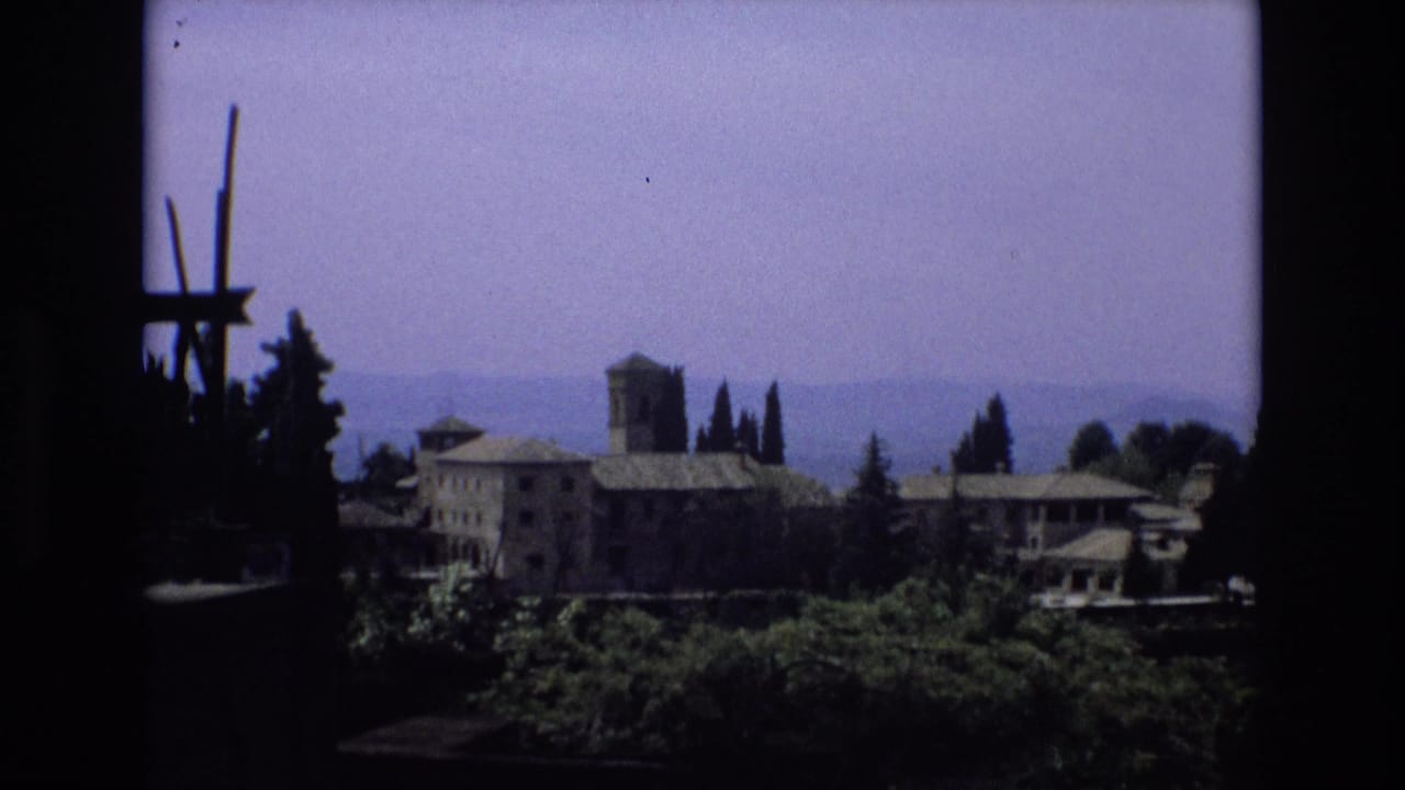 1972 Spain 8mm frame showing a distant old stone building complex with a tower, trees, hazy hills, and a dark shaded edge in the foreground.