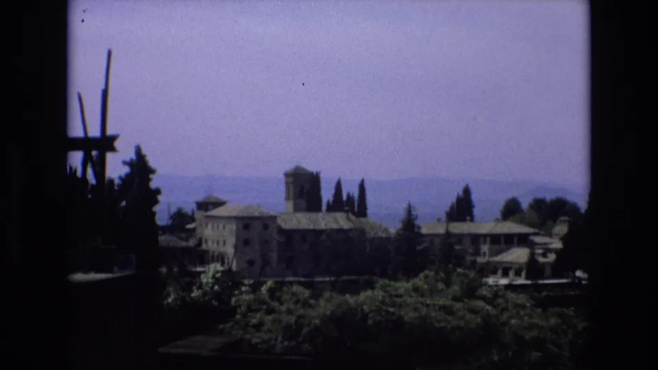 1972 Spain 8mm frame showing a distant old stone building complex with a tower, trees, hazy hills, and a dark shaded edge in the foreground.